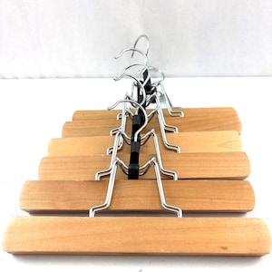 May include: A set of wooden clothes hangers with a natural finish. Each hanger features a chrome-colored metal hook and a black rubber grip. The hangers are designed for pants or skirts and are arranged in a row.