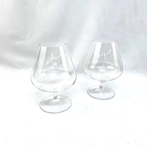 May include: Two clear brandy snifters with a wide bowl and a short stem. The glasses have a subtle etched design near the bowl. The background is a plain white surface.
