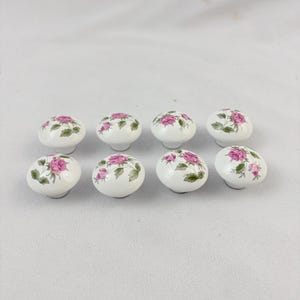 May include: A set of eight white ceramic cabinet knobs, each featuring a rounded top decorated with a painted pattern of pink roses and green leaves. The knobs are arranged in two rows of four, showcasing a smooth, glossy finish on each piece.