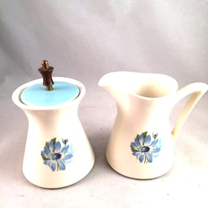 May include: Cream-colored ceramic sugar bowl with a light blue lid and a matching creamer. Both pieces feature a blue floral design. The sugar bowl has a wooden handle and the creamer has a curved handle.