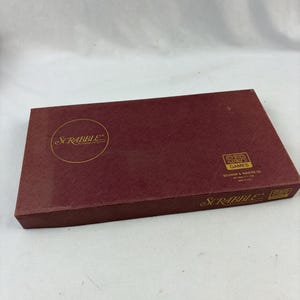 May include: A maroon Scrabble board game box. The box has gold lettering and a gold circle with the word "Scrabble" inside. The box also has the words "S&R Games" and "Selchow & Righter Co." printed on it.