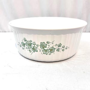 May include: White, oval-shaped serving bowl with a ribbed exterior. The bowl features a green ivy leaf pattern around the exterior. The bowl is likely made of ceramic or a similar material. The bowl is suitable for serving food.