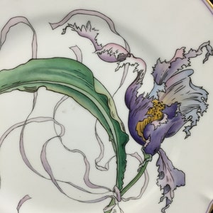 Peut inclure: Une assiette en porcelaine blanche avec un motif peint &agrave; la main d'une orchid&eacute;e violette avec des feuilles vertes et des rubans blancs.