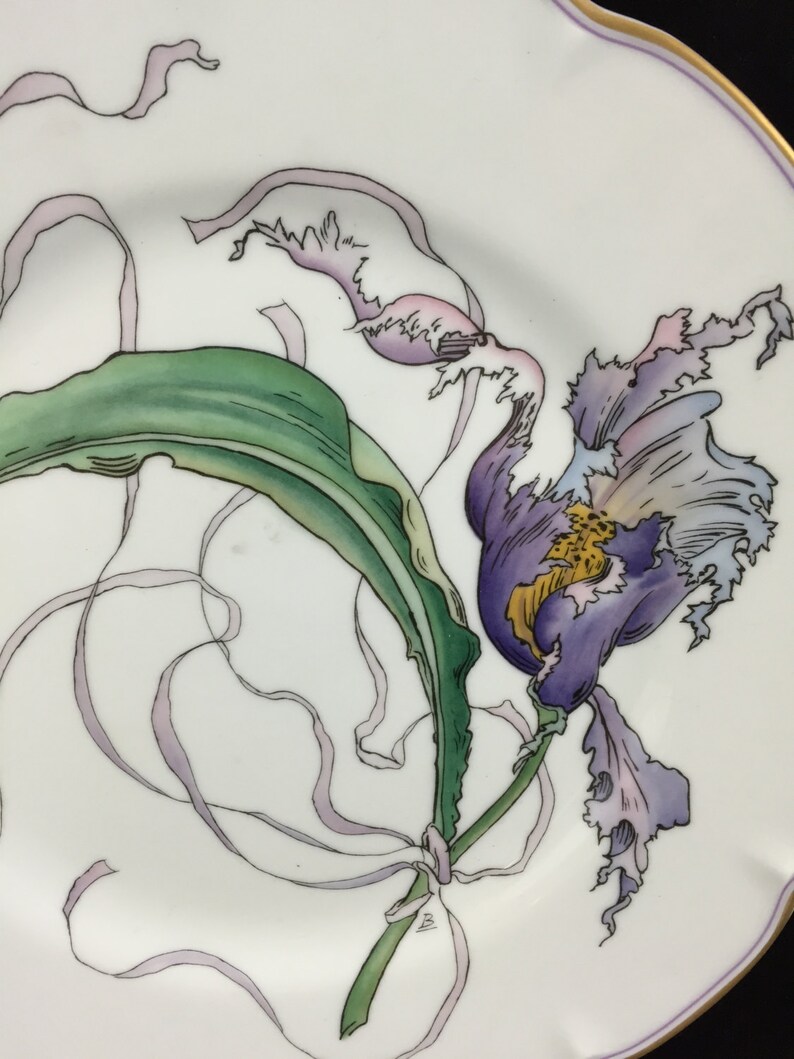 Peut inclure: Une assiette en porcelaine blanche avec un motif peint &agrave; la main d'une orchid&eacute;e violette avec des feuilles vertes et des rubans blancs.