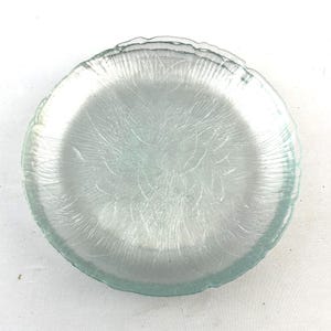 May include: A clear glass plate with a textured, etched design. The plate has a scalloped edge and a slightly bluish tint. The design features a pattern of fine lines radiating from the center.