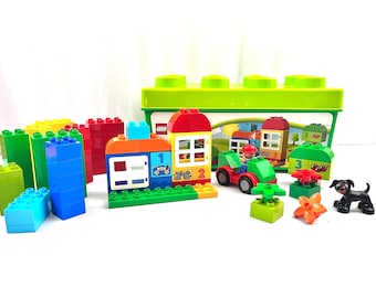 May include: A colorful Lego Duplo set featuring various building blocks, a house, a tractor with a driver, flowers, and a black dog figurine. The set includes a green storage box with a lid.