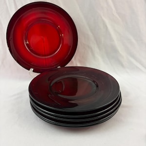 May include: A set of five circular ruby red glass saucers. One saucer is positioned upright on a small stand, while the other four are stacked horizontally. The glass features a smooth, translucent finish with a central circular indentation.