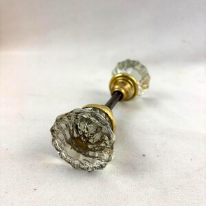 May include: A door knob set featuring two clear, faceted glass knobs with a scalloped edge design. Each knob is attached to a circular brass-toned metal base, connected by a thin, dark metal spindle rod extending between the two pieces.