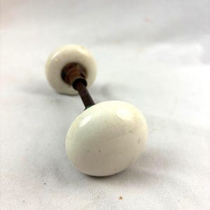 May include: A pair of round, white ceramic door knobs connected by a central metal spindle. The knobs have a smooth, glossy finish with subtle surface imperfections, and the connecting rod is made of dark, weathered metal.