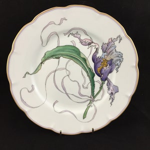 Peut inclure: Une assiette en porcelaine blanche avec un bord dor&eacute; pr&eacute;sente un motif peint &agrave; la main d'une fleur violette avec des feuilles vertes et des rubans flottants. L'assiette est parfaite pour l'affichage ou le service.