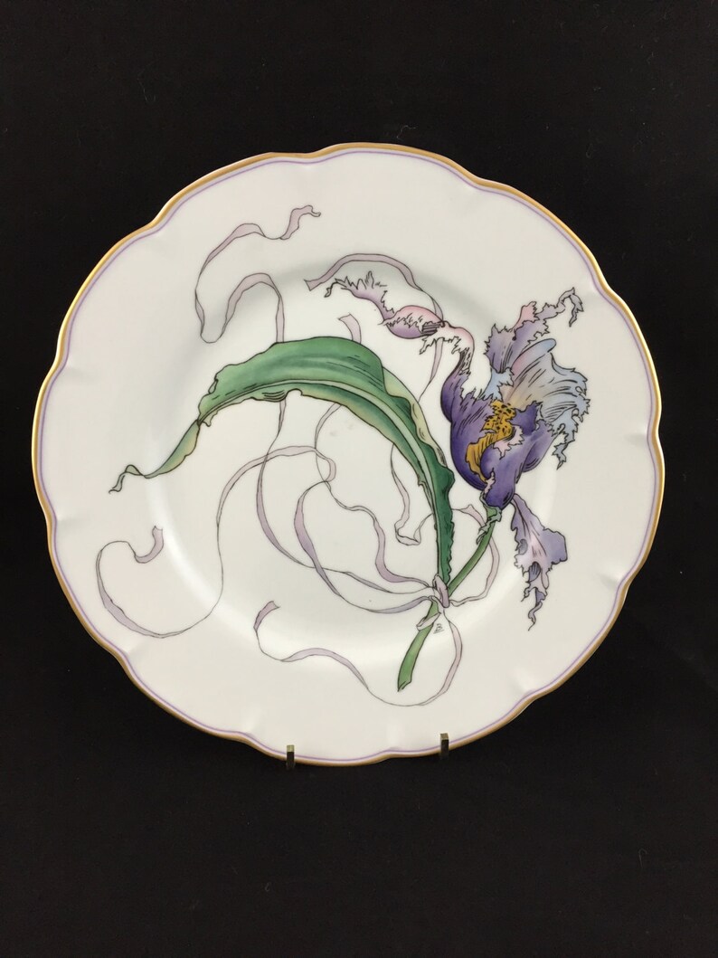 Peut inclure: Une assiette en porcelaine blanche avec un bord dor&eacute; pr&eacute;sente un motif peint &agrave; la main d'une fleur violette avec des feuilles vertes et des rubans flottants. L'assiette est parfaite pour l'affichage ou le service.