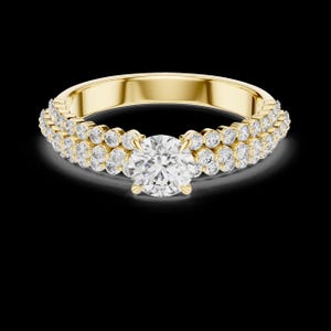 May include: A gold ring with a large, round, clear diamond center stone. The band is also adorned with two rows of smaller, clear diamonds. The ring is set against a black background.