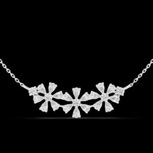 Puede incluir: Un collar de plata con tres colgantes en forma de flor, cada uno adornado con múltiples piedras preciosas transparentes. La cadena del collar es delicada y los colgantes están dispuestos en una línea curva. El fondo es negro.