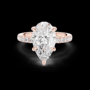 May include: A rose gold ring featuring a large pear-cut center stone held by five prongs. The band is crafted from rose gold and is adorned with a row of small, round-cut white stones set along the shank on both sides of the center pear stone.