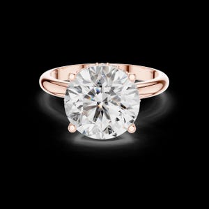 May include: Rose gold engagement ring featuring a large, round-cut diamond. The diamond is held in place by four prongs, and the band has a smooth, polished finish. The ring is set against a black background.