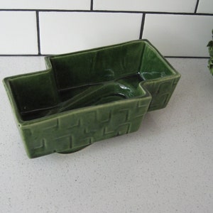 Puede incluir: Un macetero de cerámica verde con forma de camión pequeño. El macetero tiene una superficie texturizada y una pequeña abertura en la parte posterior para el drenaje.