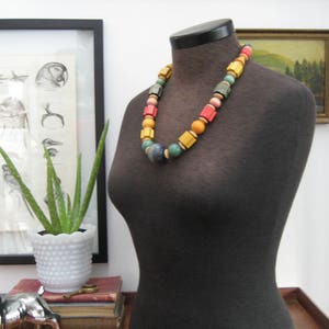 May include: A black fabric mannequin with a colorful beaded necklace. The necklace has yellow, orange, red, green, and blue beads. The mannequin is on a wooden table.