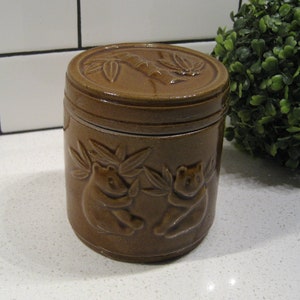 Op de afbeelding: Een bruine keramische pot met een deksel met een bamboe ontwerp en twee beren. De pot heeft een rustieke, vintage uitstraling.