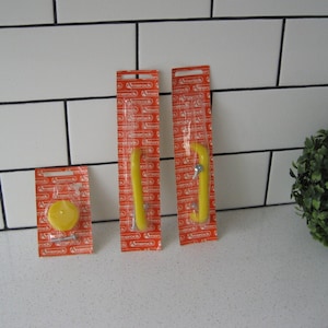 May include: Three yellow cabinet hardware pieces in their original packaging. The packaging is orange with red text. The hardware includes two handles and a knob. The items are displayed on a white surface with a white tile background.