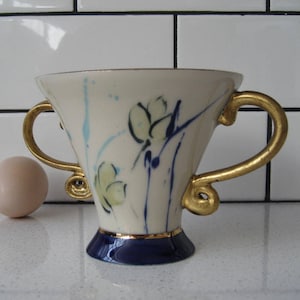 Puede incluir: Una taza de cerámica blanca con asas doradas y diseños florales azules y verdes. La taza tiene un borde y una base azules.