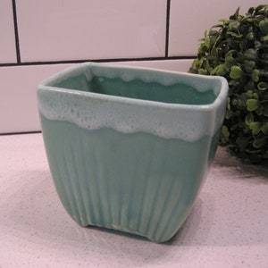 Peut inclure: Un pot carré en céramique turquoise avec un extérieur nervuré et un bord blanc texturé. Le pot est présenté à côté d'une topiaire verte et sphérique sur un fond carrelé blanc.