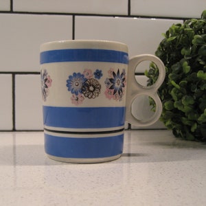 Può includere: Una tazza blu e bianca con un motivo floreale e due manici. La tazza è appoggiata su un piano di lavoro bianco.