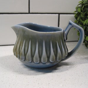 Puede incluir: Una jarra de crema de cerámica azul y verde con un diseño de lágrima. La jarra tiene un asa y un cuerpo redondeado.