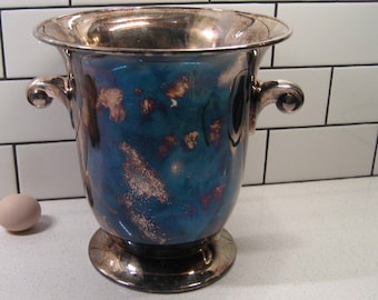 Puede incluir: Un cubo de vino plateado con un vibrante acabado azul y dorado. El cubo tiene dos asas y una base de pedestal. Un huevo está a la izquierda del cubo. El fondo es una pared de azulejos blancos.