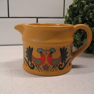 Könnte beinhalten: Ein gelber Keramik-Milchkännchen mit einem Hahn-Design. Der Hahn ist in Rot, Blau, Grün und Schwarz bemalt. Der Milchkännchen hat einen Griff und einen Ausguss.
