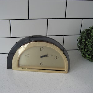 Puede incluir: Un reloj de media luna dorado y negro con agujas negras y números romanos. El reloj está sobre una encimera blanca con azulejos blancos en el fondo.