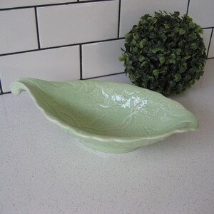 May include: A light green ceramic serving dish shaped like a leaf with a handle. The dish has a textured surface and is perfect for serving snacks or appetizers.