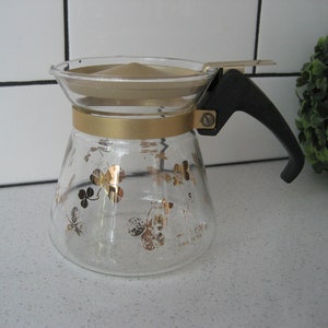 Könnte beinhalten: Eine Glas-Kaffeekanne mit schwarzem Griff und goldenen Akzenten. Die Kanne hat ein Kleeblatt-Design.
