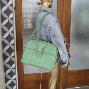 May include: A green leather travel bag with a buckle closure. The bag is being worn by a mannequin wearing a denim jacket and a brown fedora.