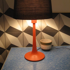 May include: A black lampshade with a red base sits on a blue table with a white and black geometric tile background. A small white ceramic pot with green and brown accents is on the table.