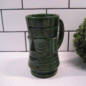 May include: A green ceramic tiki mug with a face and a handle. The mug is on a white countertop.