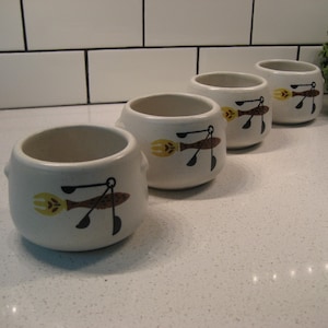 Könnte beinhalten: Vier weiße Keramik-Schalen mit braunen und gelben Designs einer Gabel, eines Löffels und eines Messers. Die Schalen sind auf einer weißen Arbeitsplatte aufgereiht.