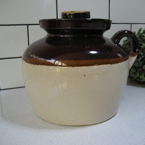 Puede incluir: Un tarro de cerámica esmaltado en marrón y blanco con tapa y asa. El tarro tiene un esmalte marrón en la mitad superior y un esmalte blanco en la mitad inferior.