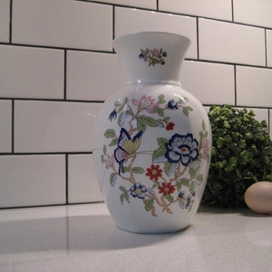 Puede incluir: Jarrón de cerámica blanco con un diseño floral que presenta mariposas, flores y hojas en tonos de rosa, azul, verde y rojo.