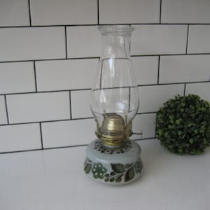 Puede incluir: Una lámpara de aceite vintage con una chimenea de vidrio transparente y una base pintada con un diseño floral en tonos verdes y azules.