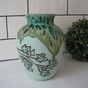 Puede incluir: Un jarrón de cerámica azul claro con un esmalte verde y marrón. El jarrón tiene un diseño de hojas pintado en la parte delantera.