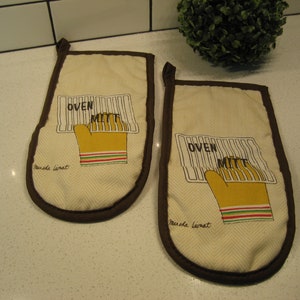 May include: A pair of beige oven mitts with brown trim. The mitts have a white chevron pattern and a graphic of a hand holding an oven mitt with the text "OVEN MITT" printed on it. The text "Mariska Linnat" is printed on the bottom of each mitt.