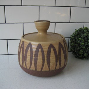 Puede incluir: Un tarro de cerámica marrón y beige con tapa y diseño geométrico. El tarro tiene una base marrón y una parte superior beige con formas de hojas marrones.