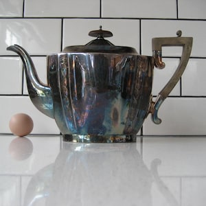 以下が含まれることがあります： 蓋に暗い木製のノブが付いた、フルーテッドデザインの銀製のティーポット。ティーポットは銀製のハンドルと注ぎ口が付いています。ティーポットは白いタイル張りの表面の上に置かれています。