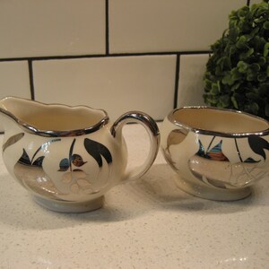 Könnte beinhalten: Ein weißes Sahne- und Zuckergefäß-Set mit silbernen Blattakzenten. Das Set zeigt ein florales Design mit Blättern und Beeren.