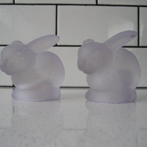 Op de afbeelding: Twee frosted glazen konijnenbeeldjes in een lichtpaarse kleur. De konijnen zitten op een wit oppervlak.