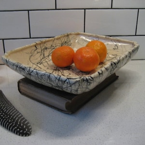 Op de afbeelding: Een witte keramische kom met zwarte draaiende lijnen, gevuld met vier oranje mandarijnen. De kom staat op een bruin boek.
