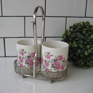 May include: A silver metal caddy with two white ceramic cups featuring pink floral designs. The caddy has a handle and is placed on a white countertop.