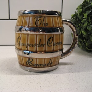 Puede incluir: Una taza de cerámica marrón con forma de barril con un borde y un asa plateados. La taza tiene el texto "The Gunpowder Cavern Bermuda" impreso en ella.