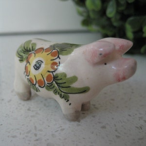 Könnte beinhalten: Eine kleine weiße Keramik-Schweinefigur mit grünen und gelben Blumendesigns. Das Schwein hat eine rosa Schnauze und Ohren.
