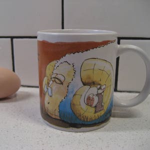 Könnte beinhalten: Eine weiße Keramiktasse mit einer Cartoon-Illustration eines Schafs mit Brille und Hut. Das Schaf sitzt auf einem Bett aus Gras und Blumen. Die Tasse hat einen braunen Henkel und einen braunen Rand.